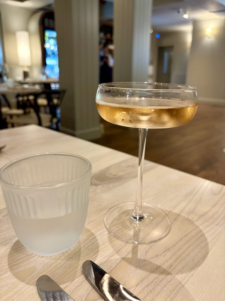 Coupe of sparkling wine next to a frosted water tumbler on a pale wooden table