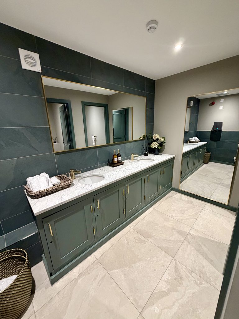Toilets with a marble double vanity, dark green tiles, rolled hand towels and a full-length mirror