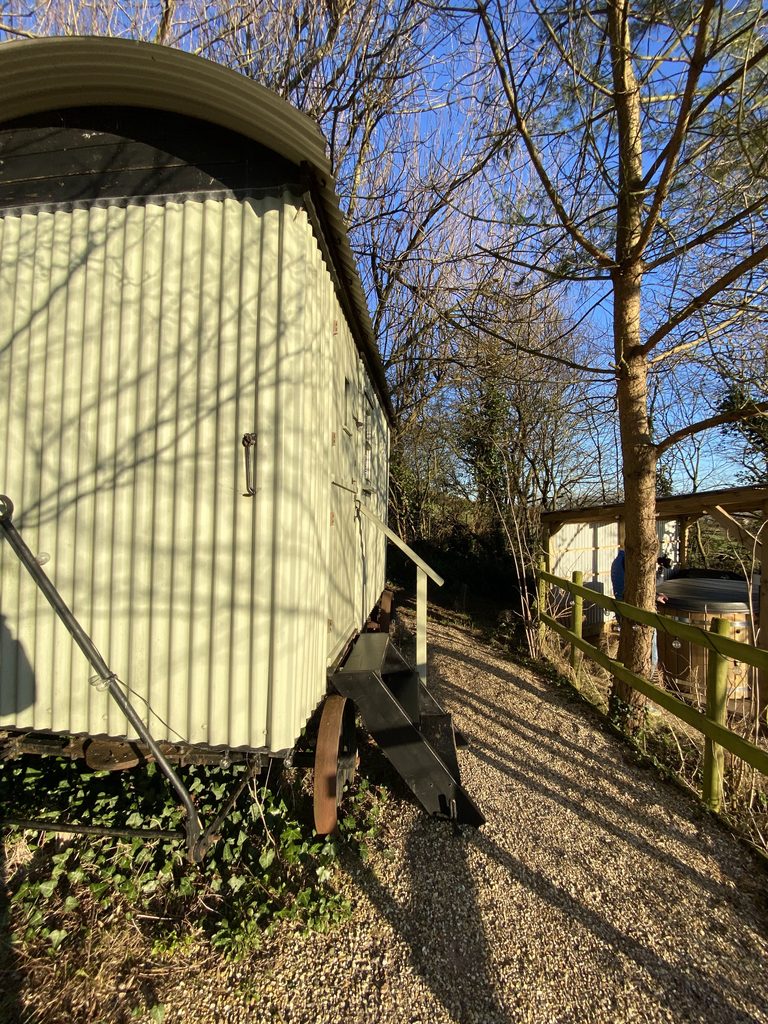 Side of the green corrugated shepherd's hut on iron wheels, with a gravel path to the hot tub shelter
