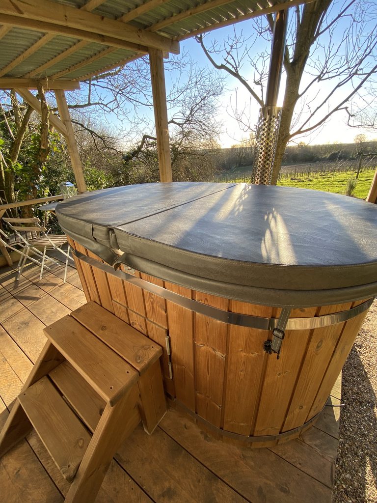Wood-fired hot tub with its cover on, under a timber shelter with steps, looking out over the vines