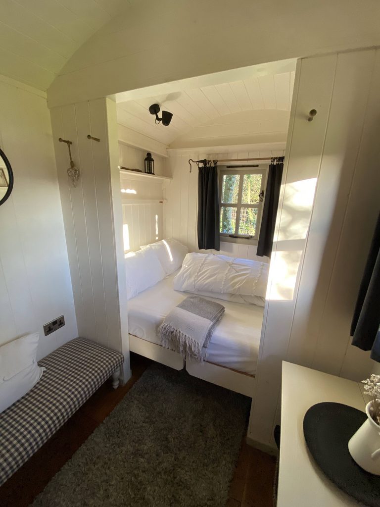 Inside the shepherd's hut: a double bed with white bedding under a window, and a checked bench seat