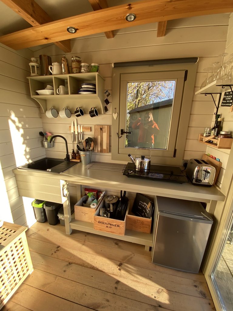 Cabin kitchenette with a sink, two-ring hob, kettle, toaster, small fridge, mugs and wooden crates of pans