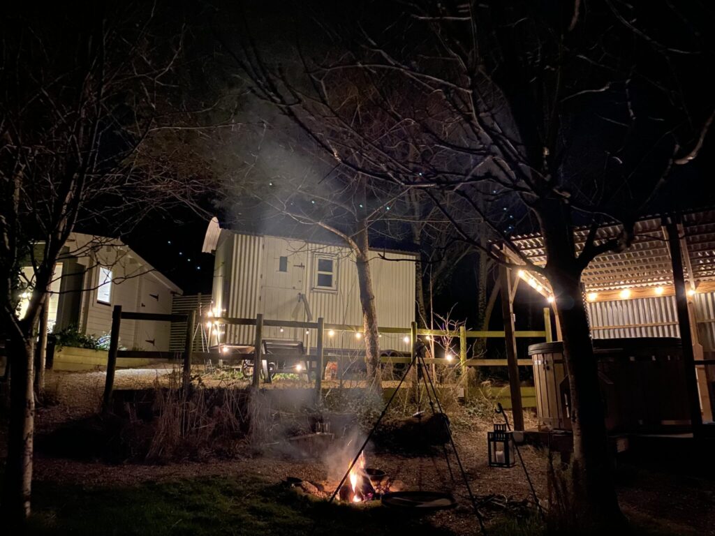 Little Waddon Vineyard shepherd's hut at night, with the cabin, the hot tub shelter and the fire pit lit