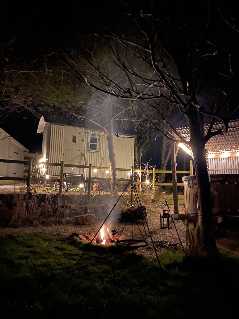 The fire pit burning at night, with festoon lights along the fence and the shepherd's hut beyond