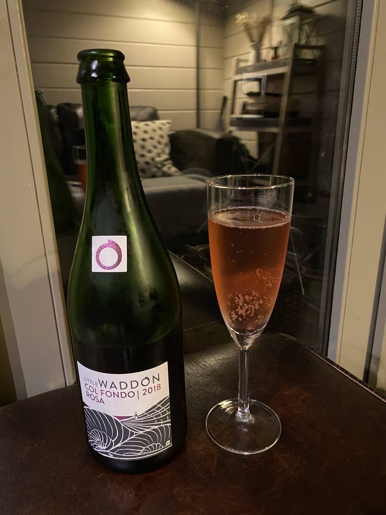 Bottle of Little Waddon Col Fondo Rosa 2018 beside a glass of pink sparkling wine, by the cabin window