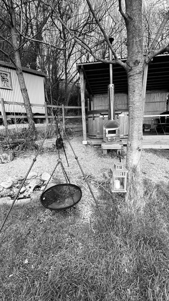 Black and white view of the fire pit with a hanging grill, the wood-fired hot tub and the hut behind