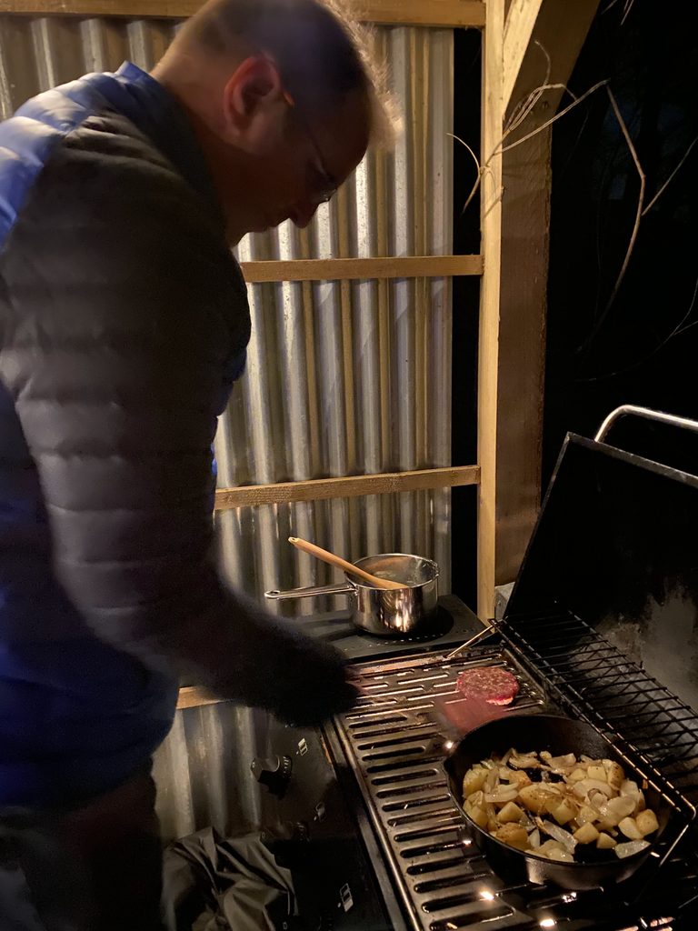 A man cooks a burger and a pan of potatoes and onions on the gas BBQ under the shelter at night