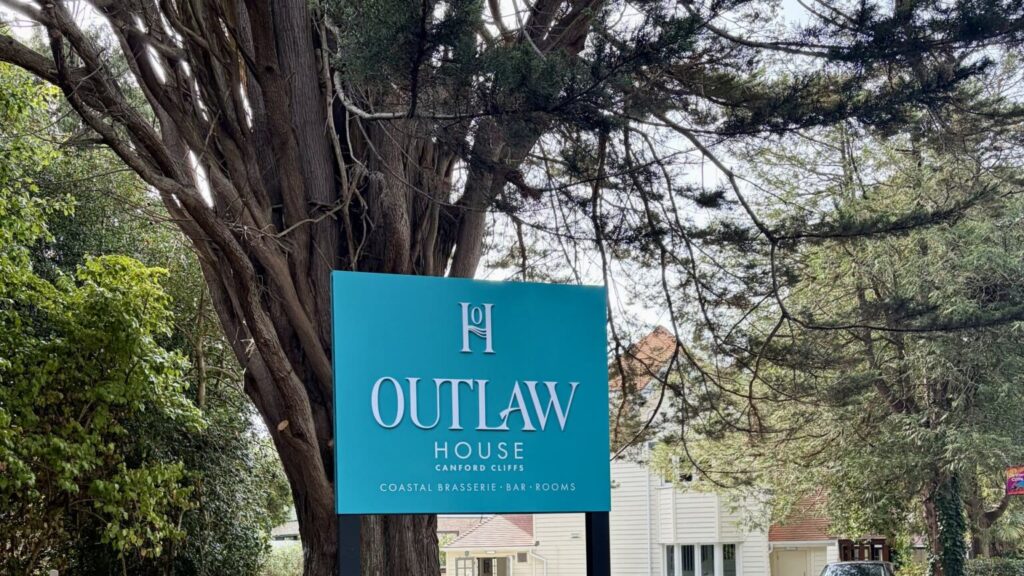 Teal Outlaw House Canford Cliffs sign under a large tree, with the white building behind